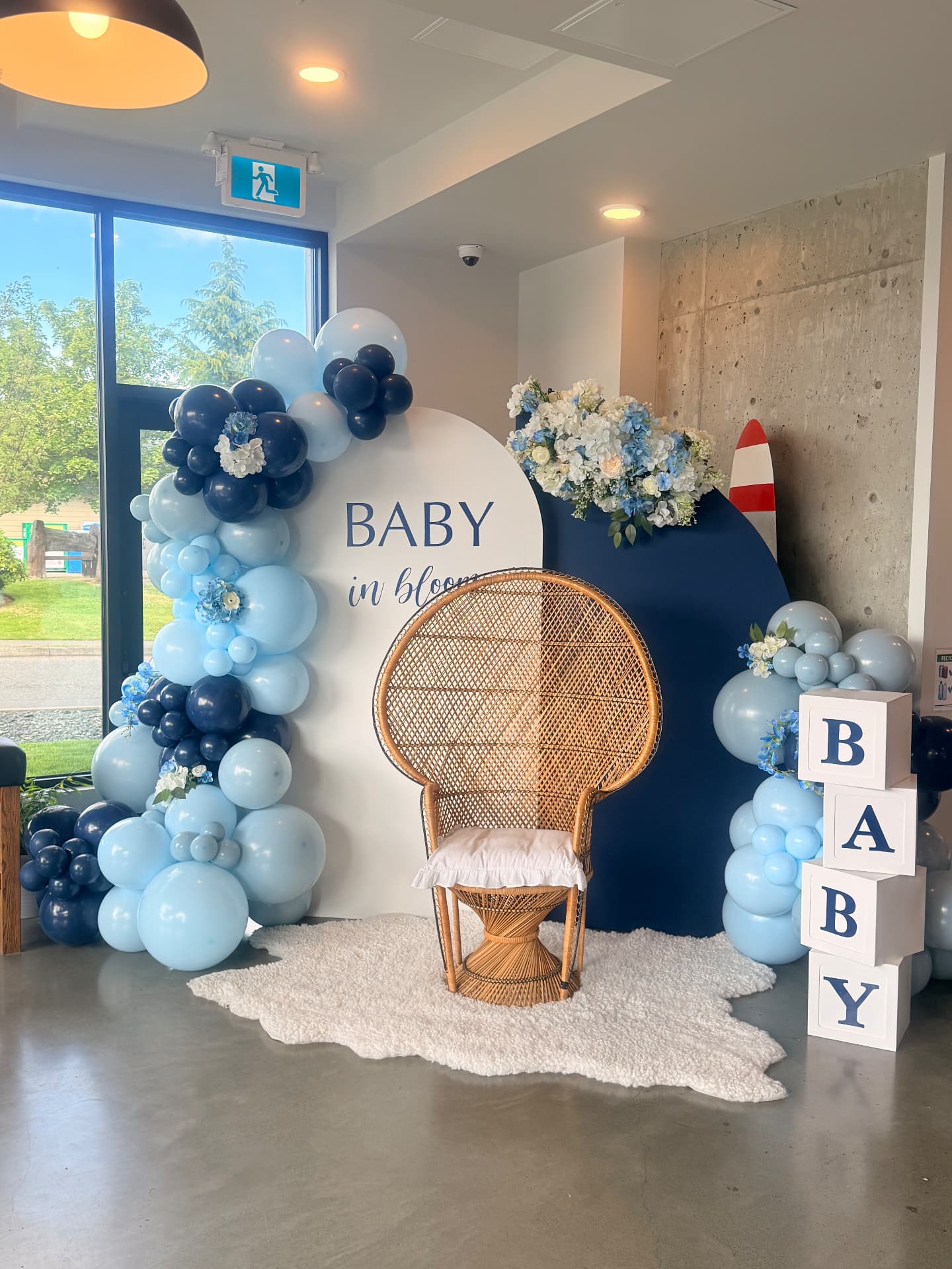 Blue baby shower backdrop with Baby in bloom sign, wicker chair and balloon garland
